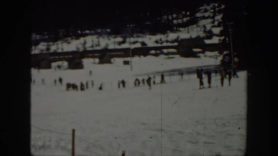 People stand near the base of a snowy recreational slope, with low wooden structures and dark trees visible in the background.