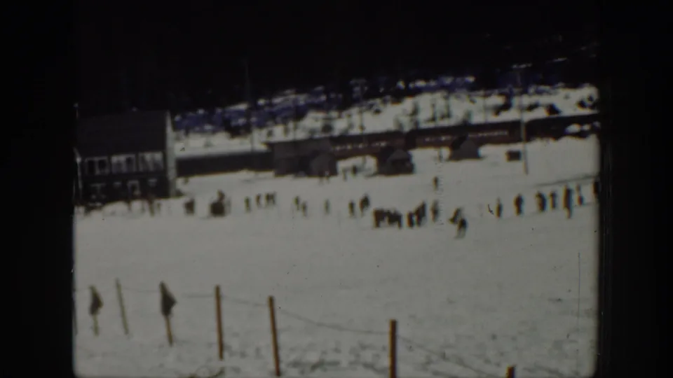 Scattered people stand and move near a snowy slope base in Washington, with fencing, posts and a lodge-like wooden structure behind them.