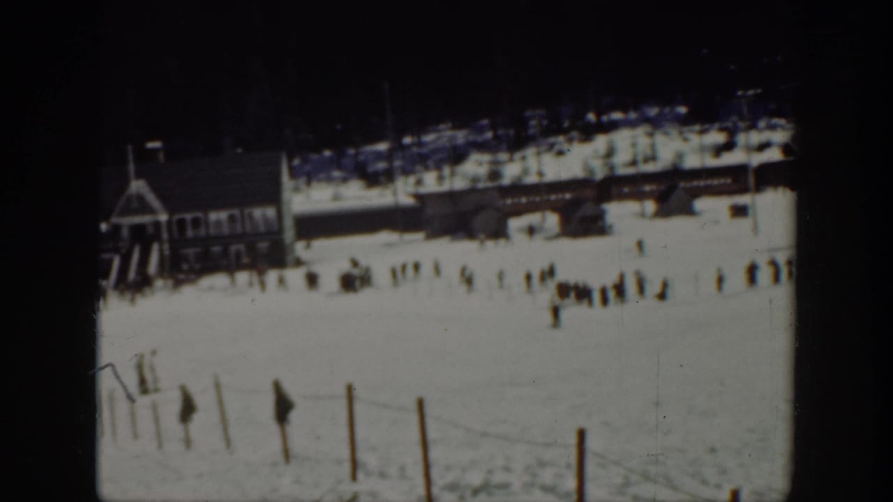 1940 Washington 8mm frame showing a snowy ski area with a wooden lodge, distant skiers, fence posts, and dark forest beyond.