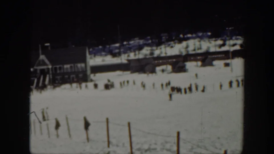 1940 Washington 8mm frame showing a snowy ski area with a wooden lodge, distant skiers, fence posts, and dark forest beyond.