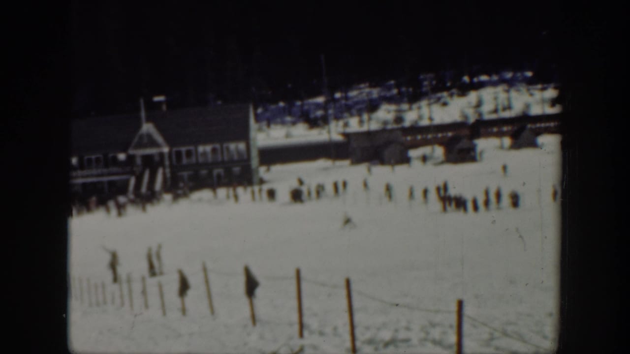 Distant skiers cross a snowy slope beside a lodge and foreground fence posts.