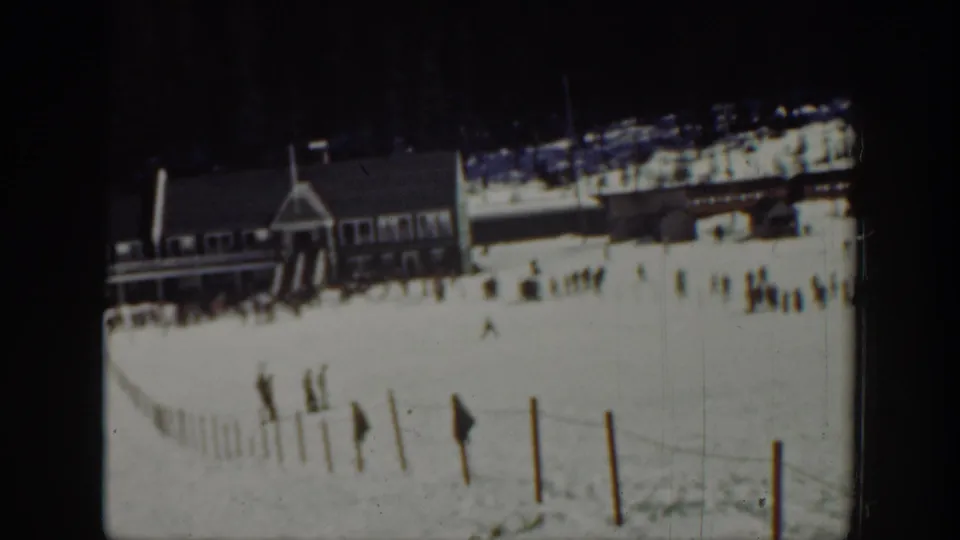 1940 Washington 8mm frame showing skiers walking uphill near slope fencing, with a wooden lodge and gathered people in the distance.