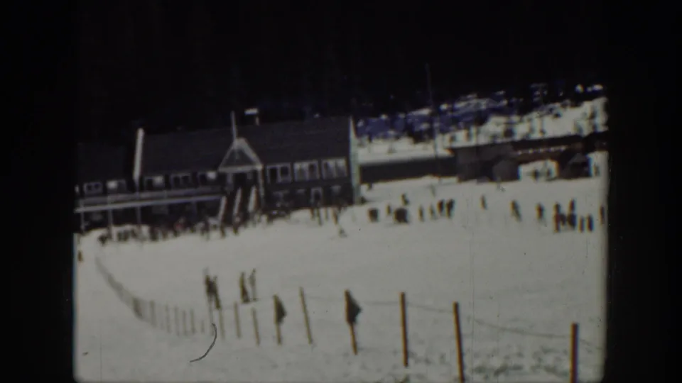 1940 Washington 8mm frame showing a ski-area lodge, scattered skiers, a snowy foreground fence, and dark trees behind the slope.