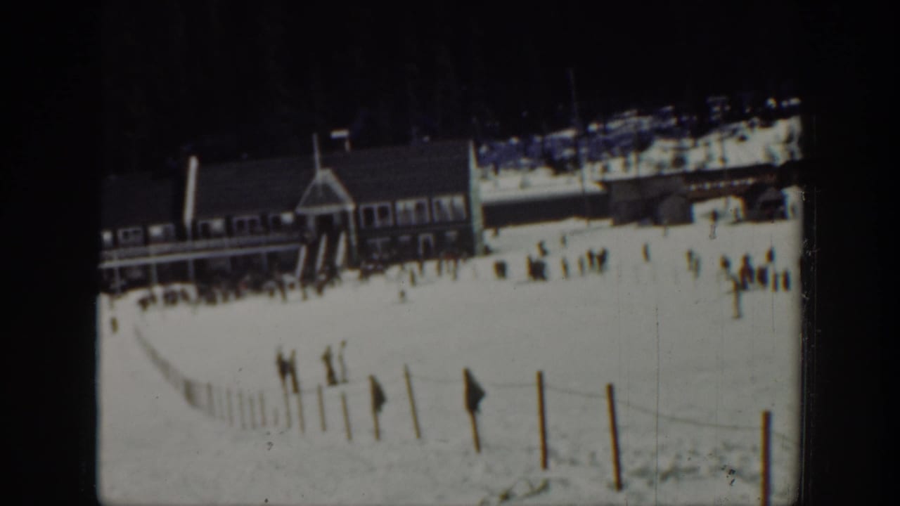 Washington 1940 home-movie frame showing a snowy ski area with a wooden lodge, fence line, and scattered visitors near the base of a slope.