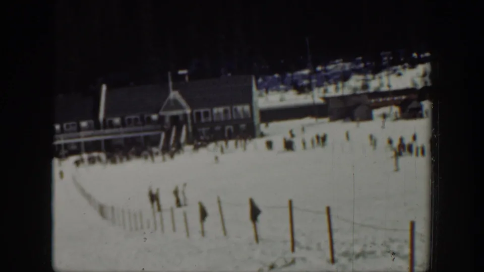 Washington 1940 home-movie frame showing a snowy ski area with a wooden lodge, fence line, and scattered visitors near the base of a slope.