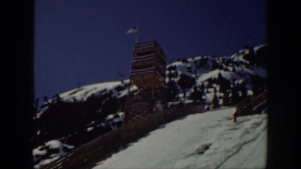 Washington 1940 frame showing a wooden observation tower with an American flag on a snowy ridge above trees and ski terrain.