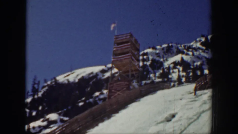 Washington 1940 home-movie frame of a wooden tower with an American flag, snowy hillside, trees, and clear blue sky.