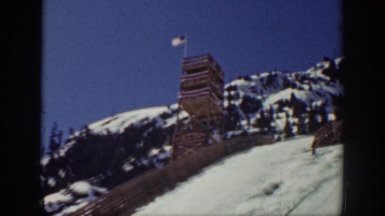 1940 Washington home-movie frame showing a snowy ski slope, wooden jump tower, American flag, rocky ridge, and a skier moving near the ramp.