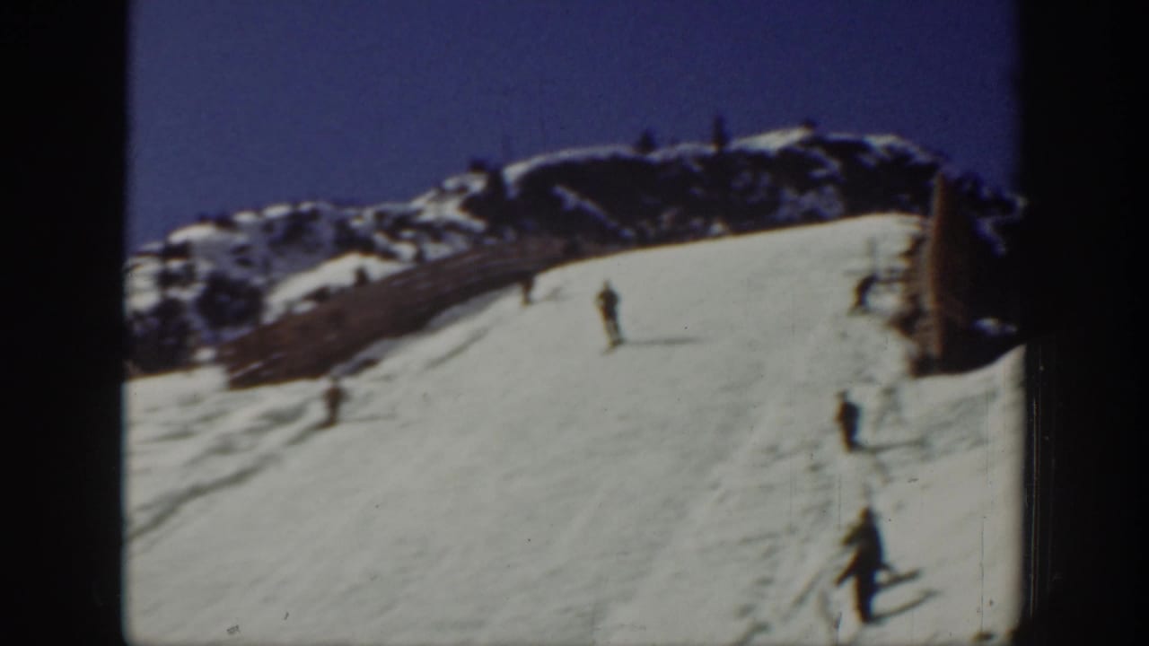 Wide snowy slope with several skiers descending below a rocky ridge and blue sky.