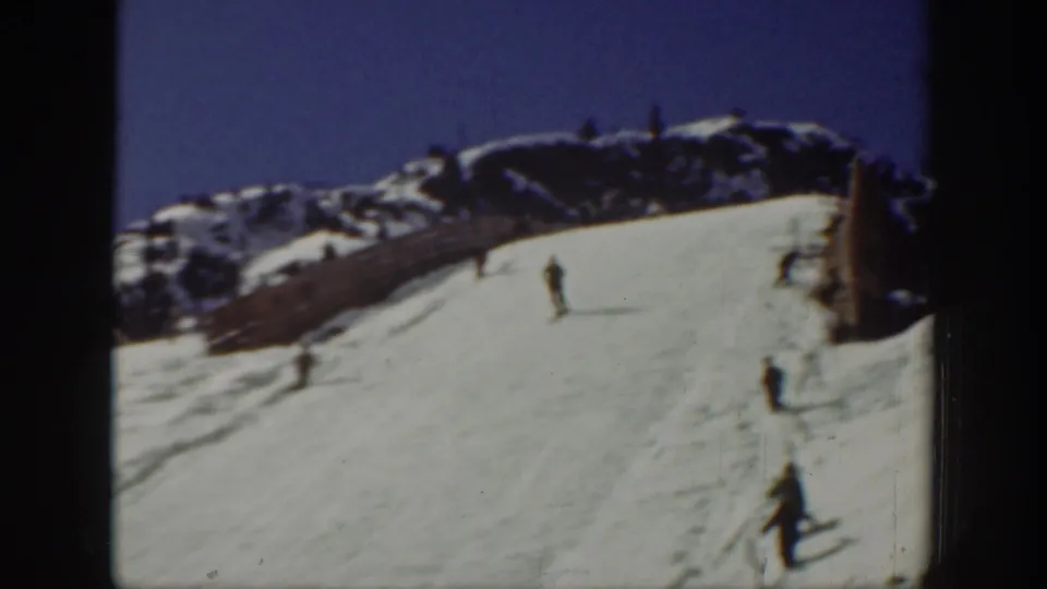 1940 Washington ski scene with several distant skiers descending a bright snow slope beneath a rocky ridgeline and deep blue sky.