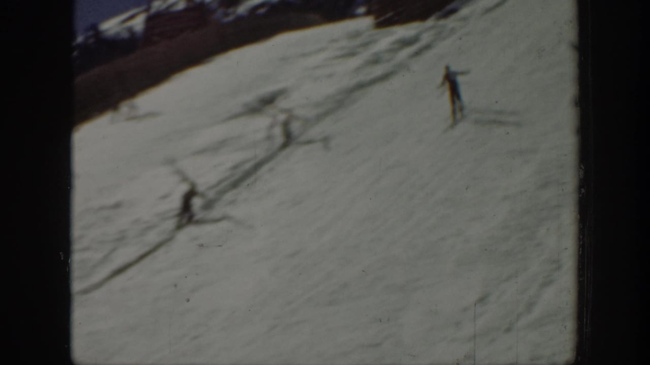 1940 Washington home-movie frame showing small skiers moving down a snowy mountain slope beneath a dark ridge and blue sky.