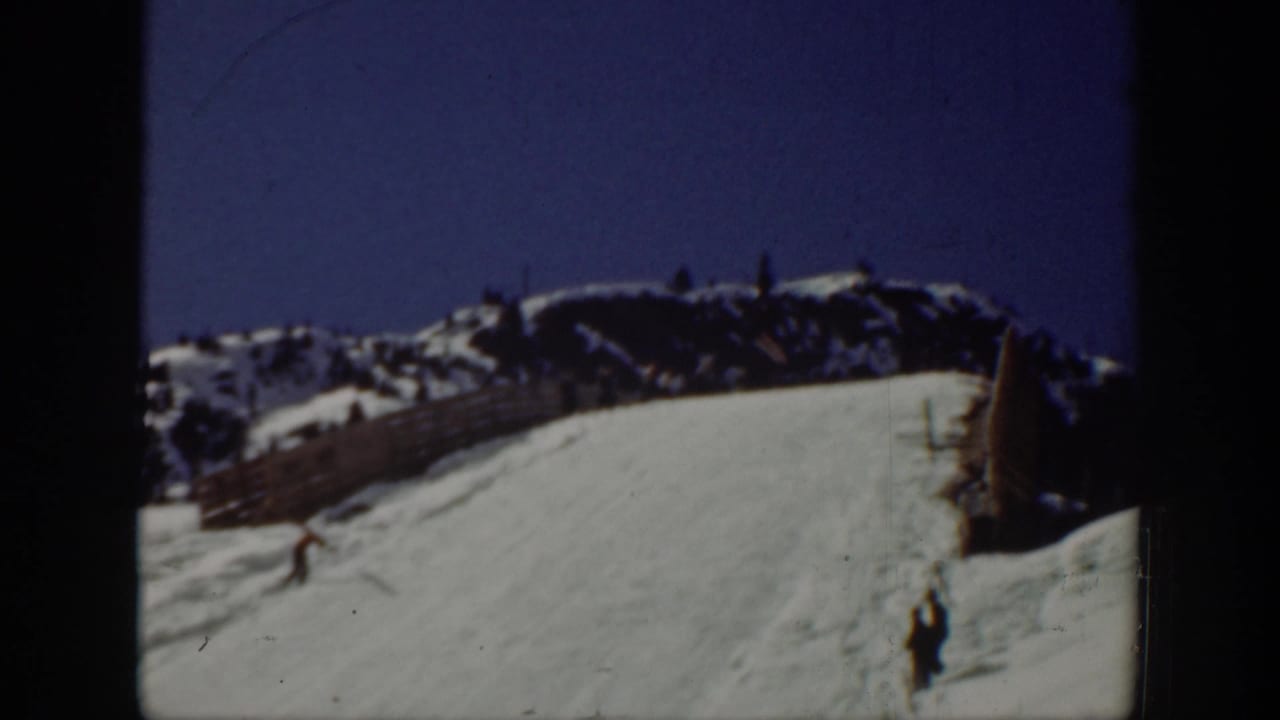 A skier moves across a snowy slope near a fence with mountains behind.