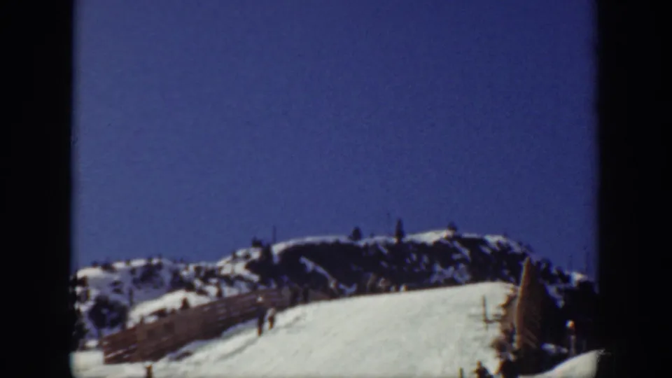 1940 Washington ski resort view with a snow-covered slope, fence line, distant people, and mountain ridge under blue sky.