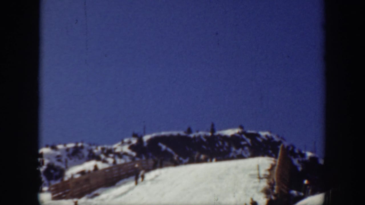 Snowy ski slope beneath a blue sky with distant ridges and small skiers on the hill.