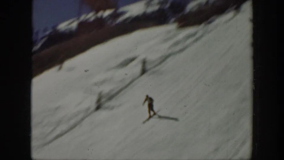 Home-movie frame from the Washington ski clip showing one skier moving downhill across a bright snowy slope with a dark mountain ridge above.