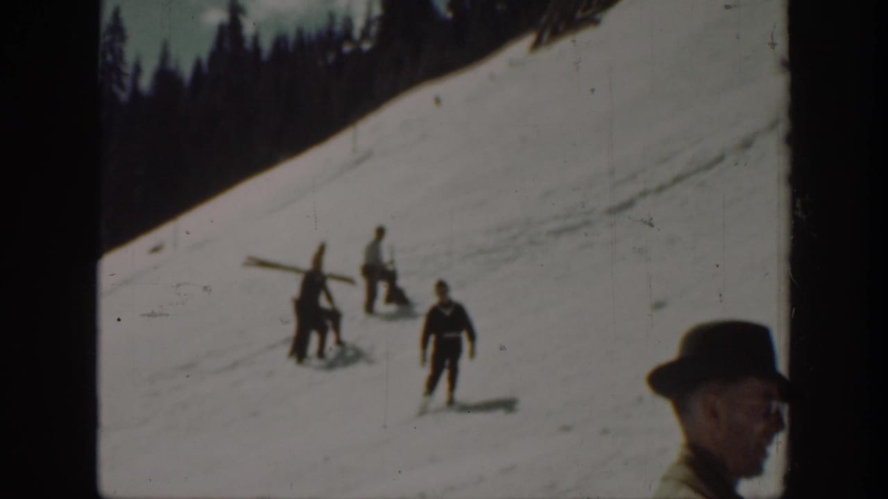 Several men stand and walk on a snowy hillside, with one carrying skis.