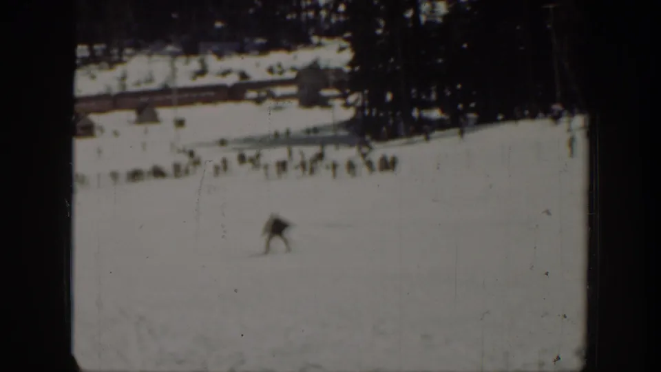 Wide 1940 Washington home-movie frame showing a skier on snow with a tree line, distant buildings, and small groups of people.
