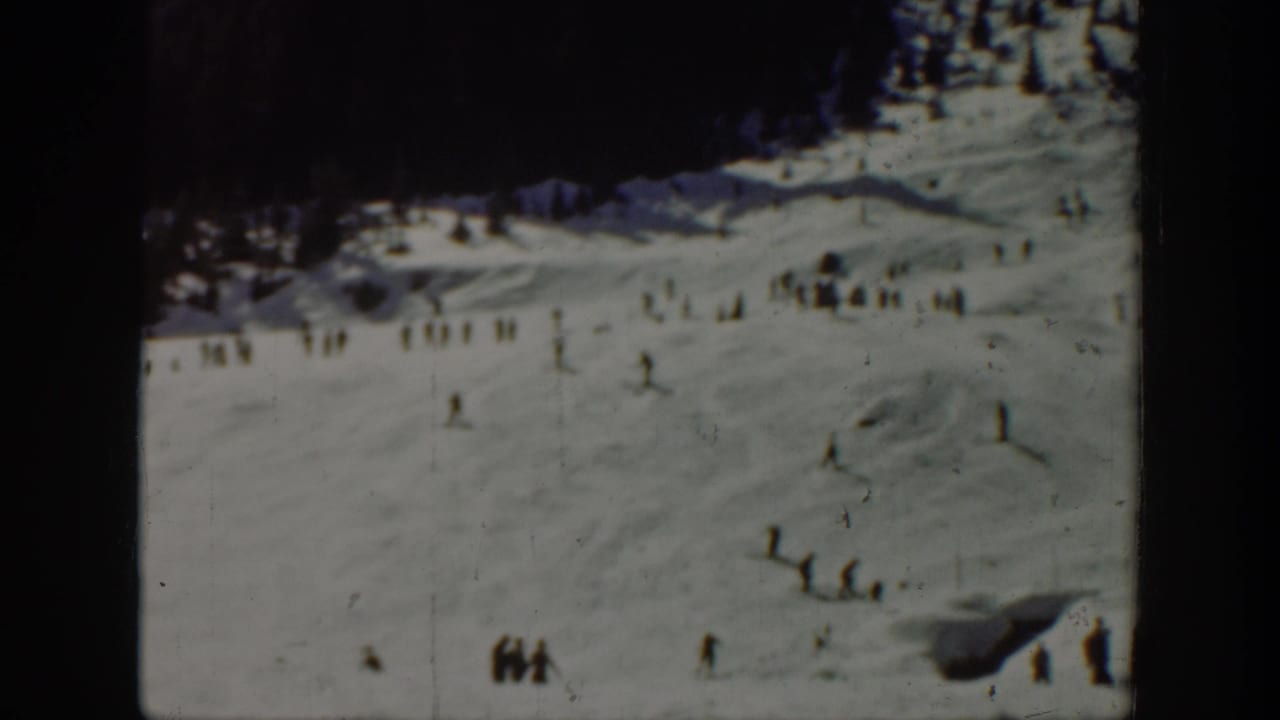 1940 Washington home-movie frame showing distant skiers and small groups on a snow-covered slope with dark trees at the upper edge.