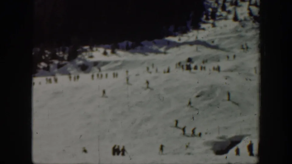 1940 Washington home-movie frame showing distant skiers and small groups on a snow-covered slope with dark trees at the upper edge.