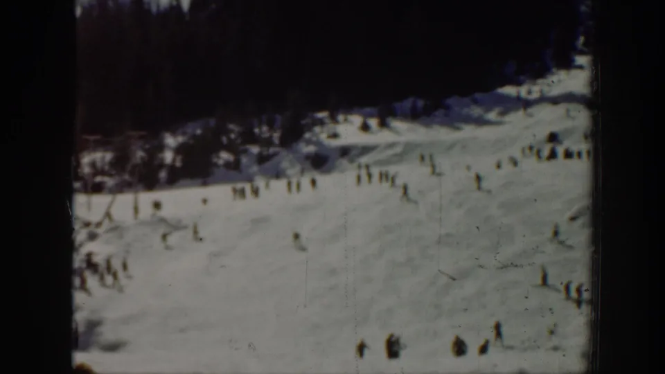 Distant 8mm view of a Washington ski resort slope with skiers, seated or standing visitors, and dark evergreen woods along the upper edge.