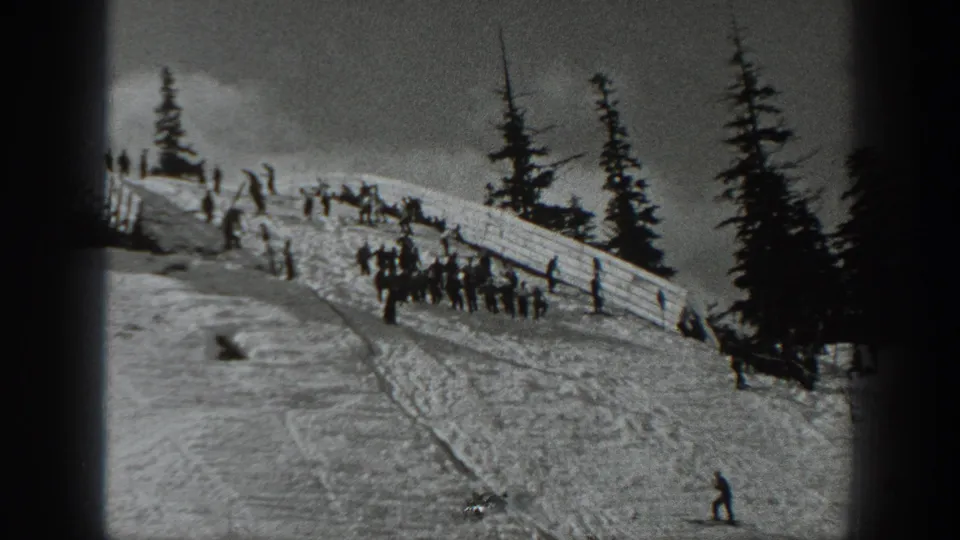 Home-movie view of skiers beside a wooden fence on a Washington snow slope, with tall evergreens framing the scene.