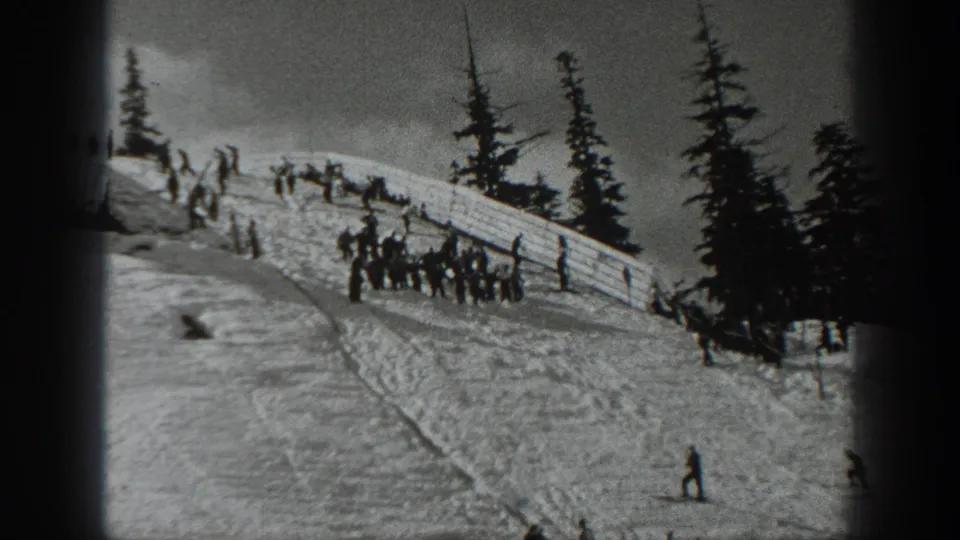 Black-and-white 1940 Washington frame with skiers and spectators gathered near a wooden fence on a snowy hill.
