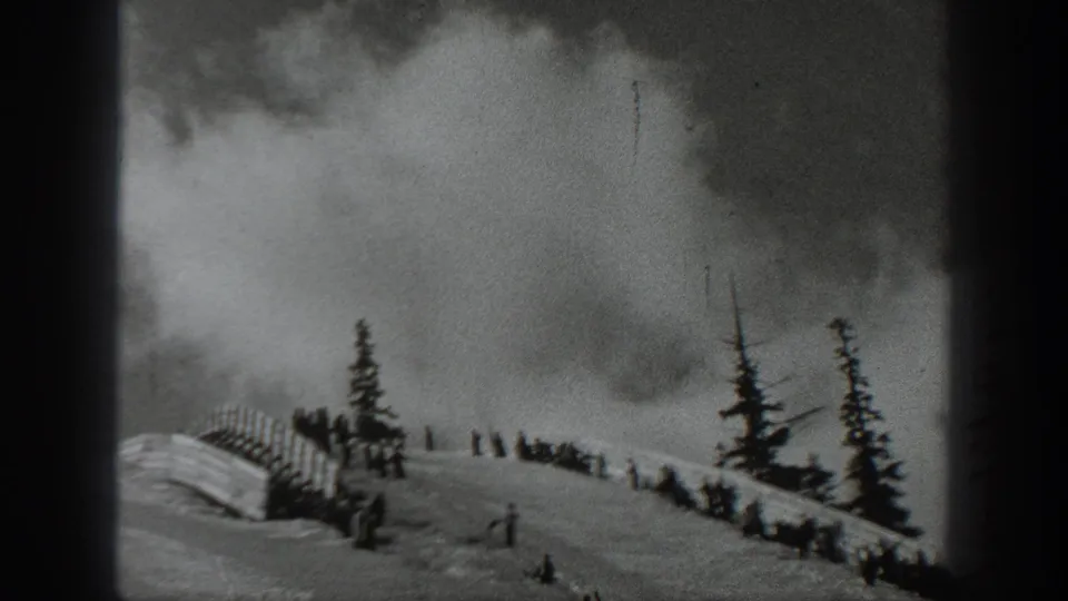 Black-and-white 8mm frame of a Washington snowy hillside with small figures near fencing under a spreading smoke cloud.