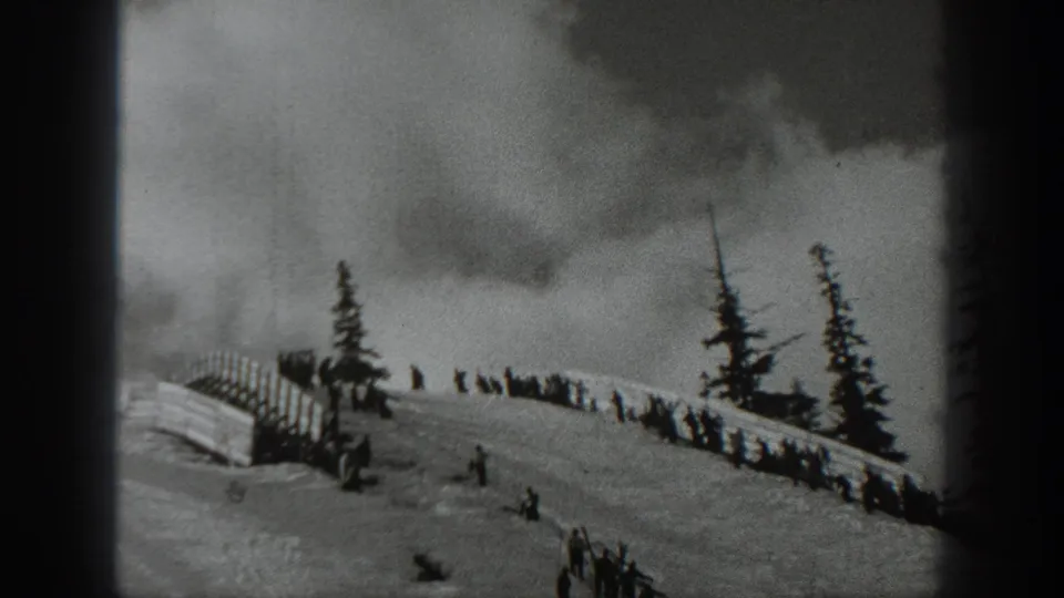 1940 Washington home-movie frame showing a snowy hillside, distant skiers, spectators by a wooden fence, evergreens, and a cloudy sky.