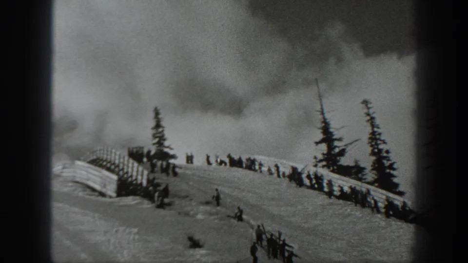 Black-and-white 8mm frame from Washington showing people gathered on a snowy ski slope with a fence, evergreens, and overcast sky.