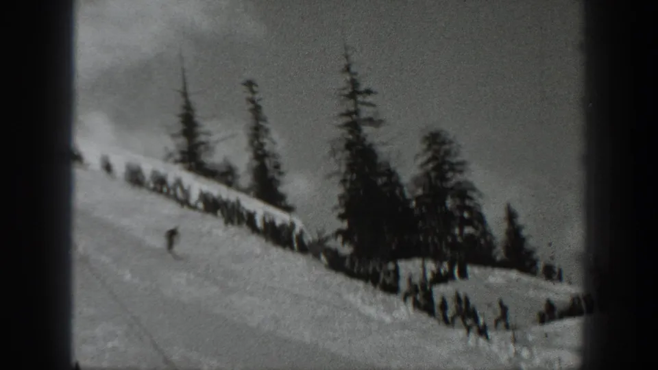 Washington ski-slope frame showing distant people near a wooden fence, a skier on the snow, tall evergreens, and black-and-white film texture.