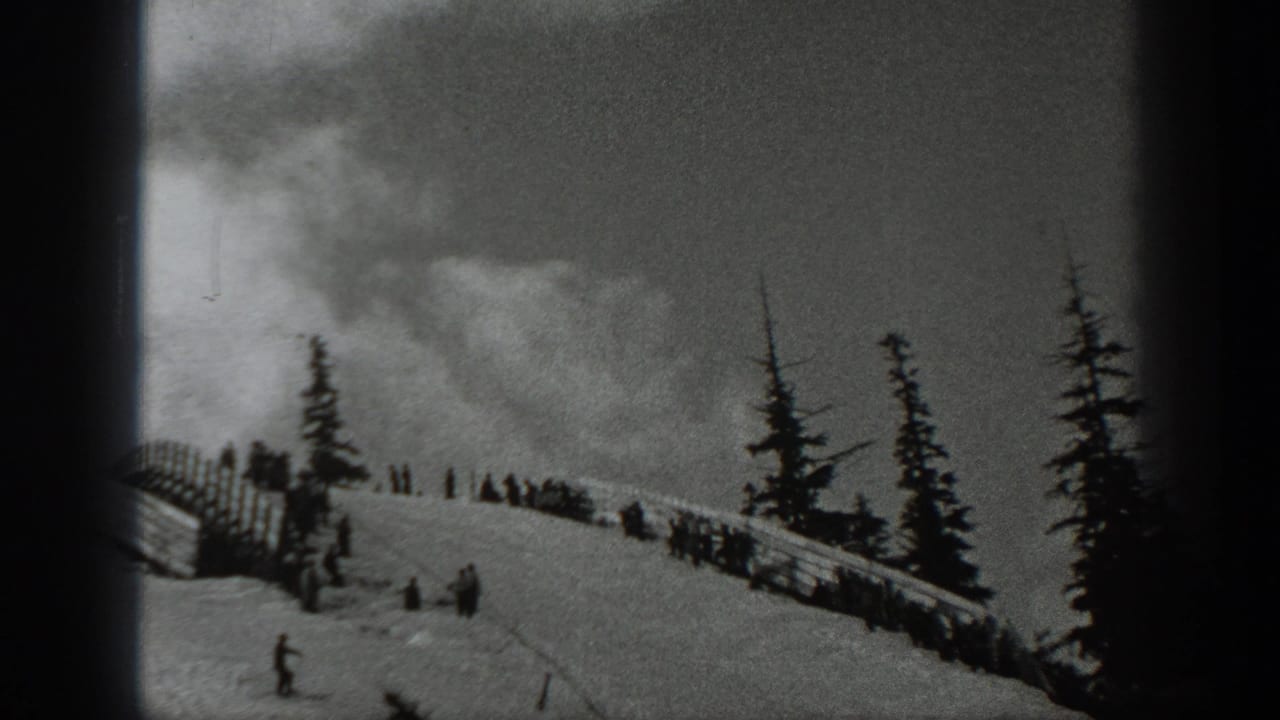 Several distant skiers stand and move on a snowy ridge near wooden fencing and tall evergreens.
