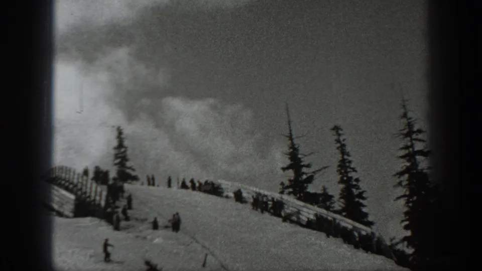 Black-and-white 1940 Washington frame with distant skiers on a snow-covered ridge near wooden snow fencing and evergreen trees.