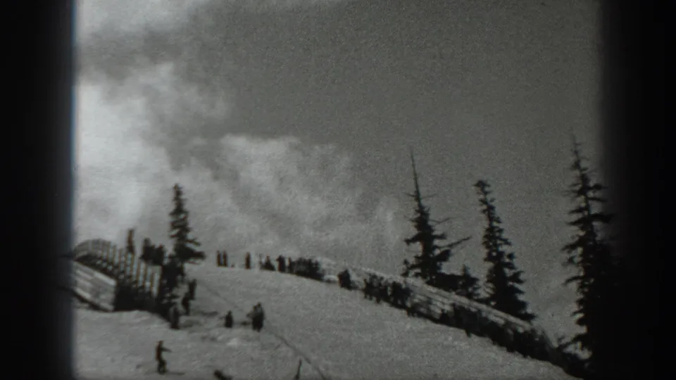 Black-and-white 1940 Washington frame showing small skiers on a snowy mountain slope with evergreens, fencing, and overcast sky.