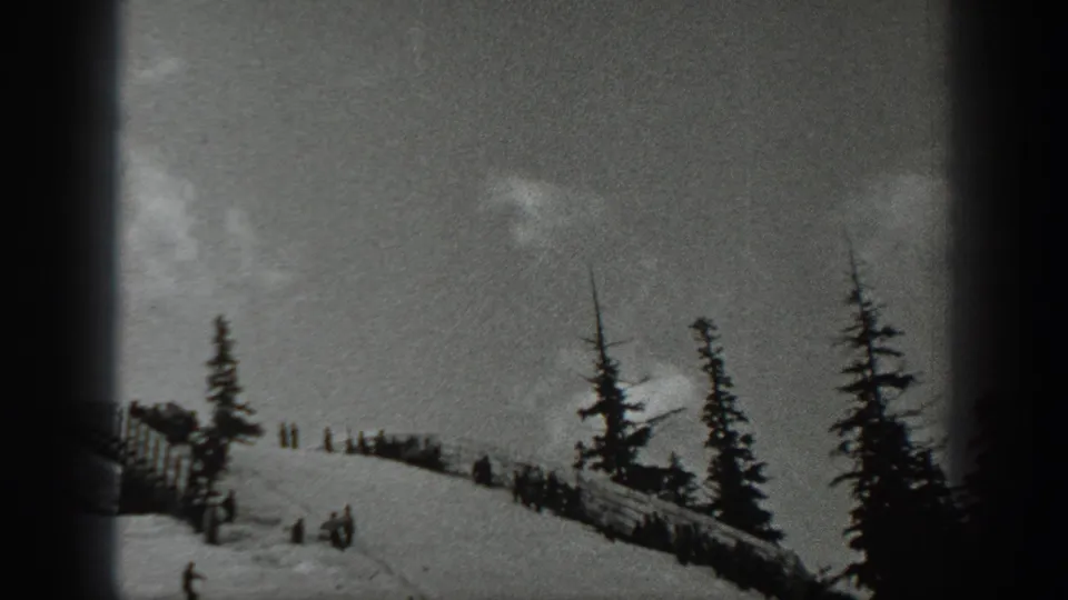 1940 Washington frame showing distant people clustered by a wooden fence on a snowy ski slope lined with evergreen trees.