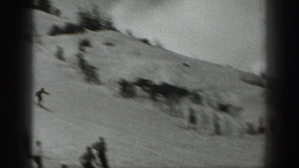 Motion-soft 8mm frame of a skier crossing a snowy hillside while scattered spectators stand near the run.