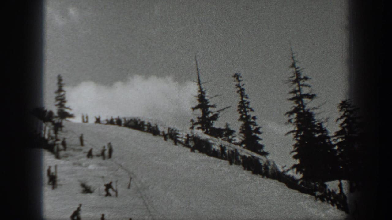 Skiers and spectators on a snowy ridge bordered by evergreen trees under a cloudy sky.
