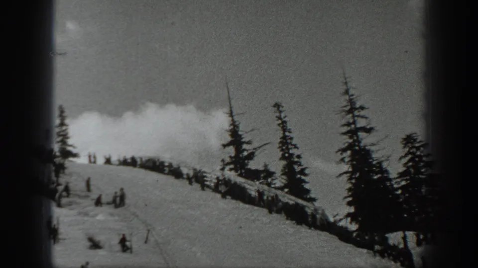 Black-and-white 8mm frame of a Washington ski slope with skiers below and people clustered near the tree line.