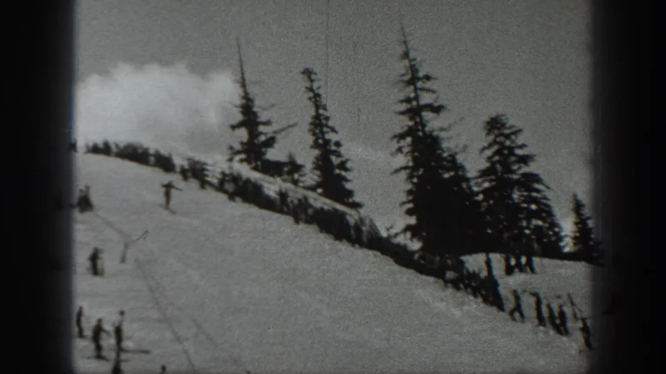 1940 Washington home-movie still showing scattered skiers on a snowy run beside tall evergreens and a cloudy ridge.
