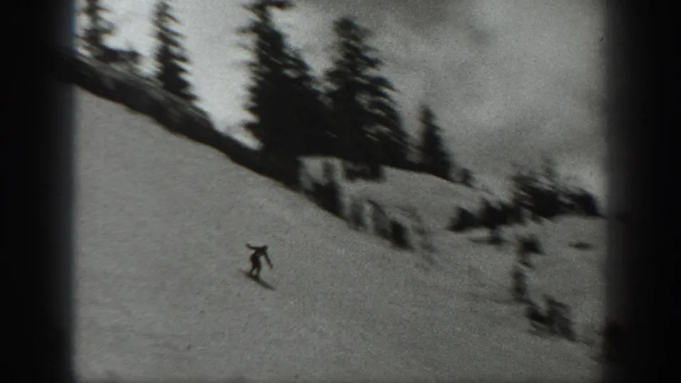 Soft, grainy 8mm frame of a skier traversing a snowy Washington hillside with blurred groups near evergreens.