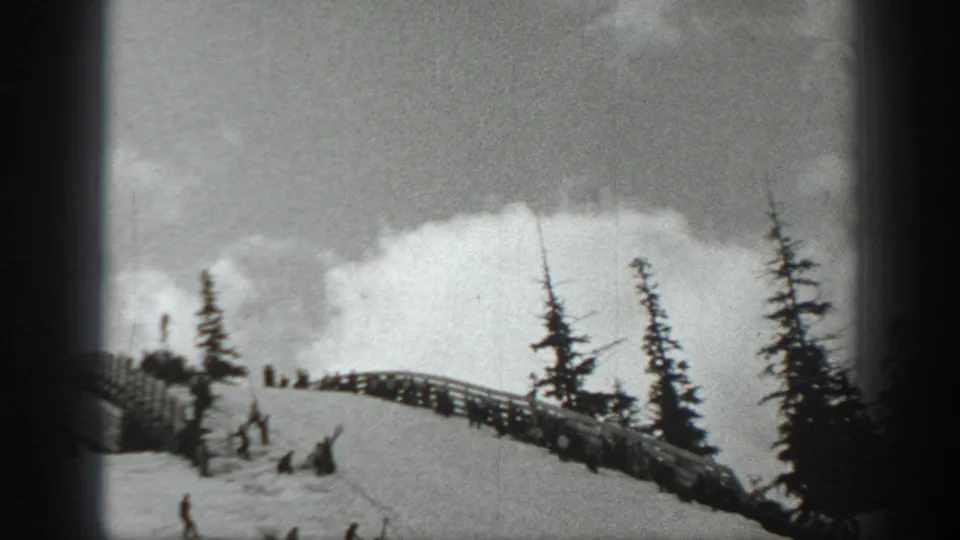 Black-and-white home-movie frame of skiers and spectators on a snowy Washington ridge under a bright cloudy sky.