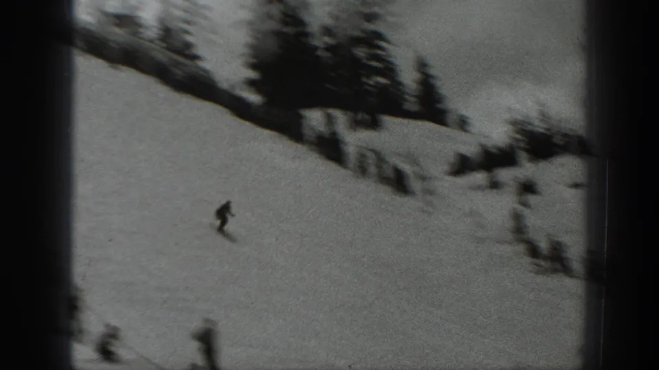 1940 Washington 8mm frame with a skier descending a snowy hillside, while blurred onlookers and evergreen trees stand near the upper slope.