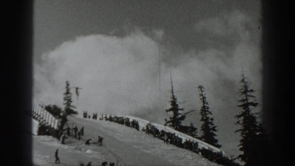 1940 Washington 8mm frame showing skiers and onlookers gathered beside a fenced snowy slope, with evergreen trees and cloudy mountain weather.