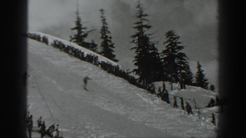 1940 Washington 8mm frame showing a skier in motion on a snowy slope, with evergreen trees, a fence line, and spectators at the side.