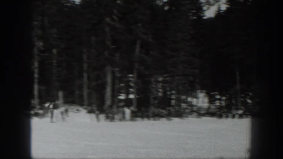 Soft black-and-white 8mm frame from Washington showing a snowy clearing bordered by dense evergreens and distant people near the treeline.