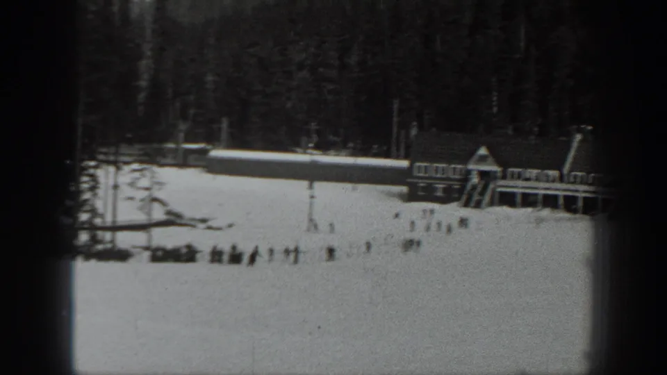 Black-and-white 1940 Washington 8mm frame showing distant figures on snow near evergreens and a rustic pitched-roof building.