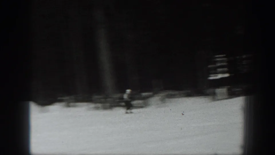 Soft black-and-white 8mm frame showing a skier moving across snow near a dark structure or tree line in Washington.
