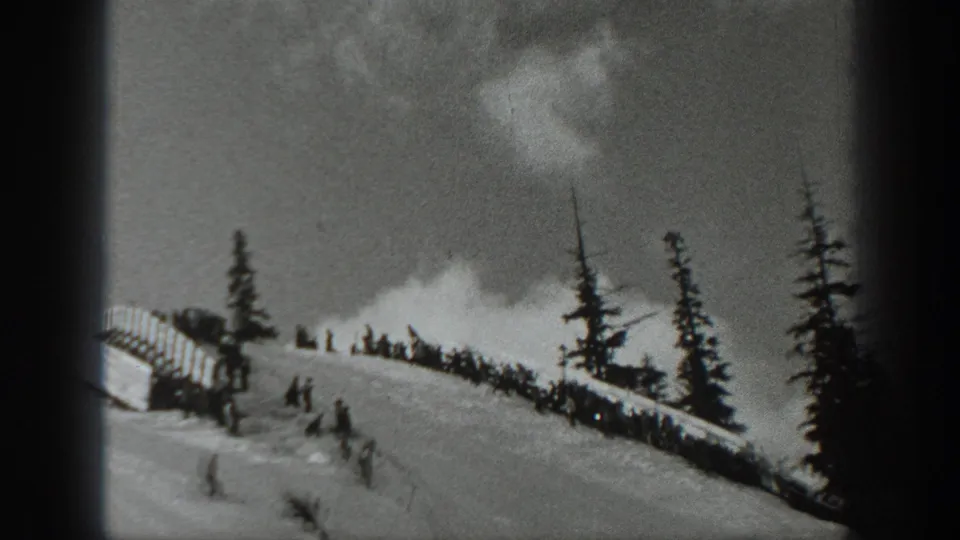 Black-and-white 1940 Washington frame showing skiers and spectators gathered near a snowy ski jump ridge under cloudy sky.