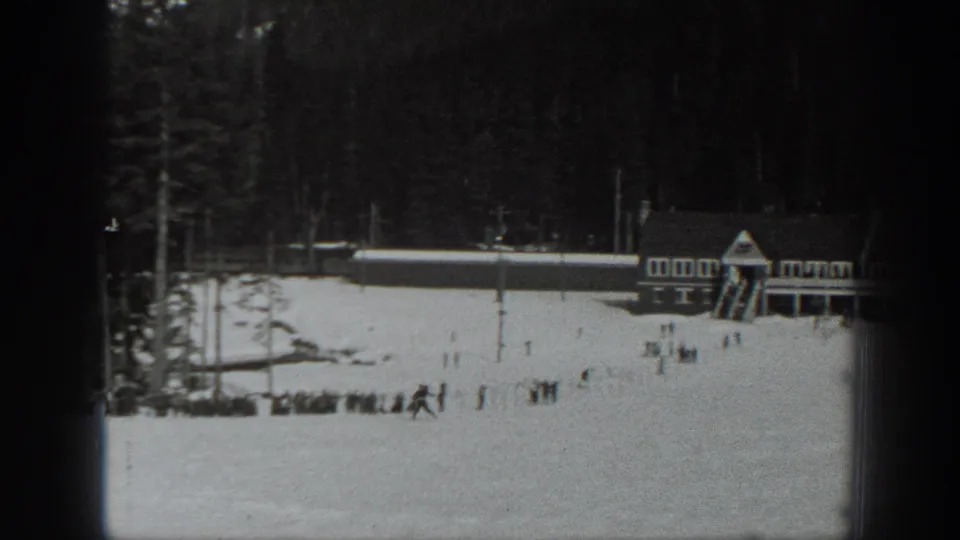 Black-and-white 8mm frame showing a lodge-like structure beside a snowy Washington field with distant visitors.