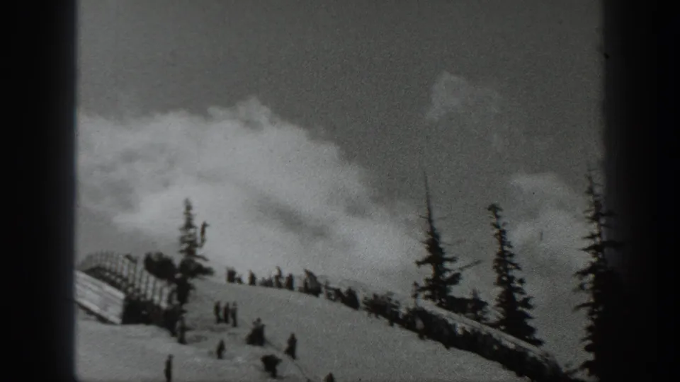 1940 Washington 8mm frame of skiers scattered across a snowy hillside with evergreen trees and a small barrier at the slope edge.