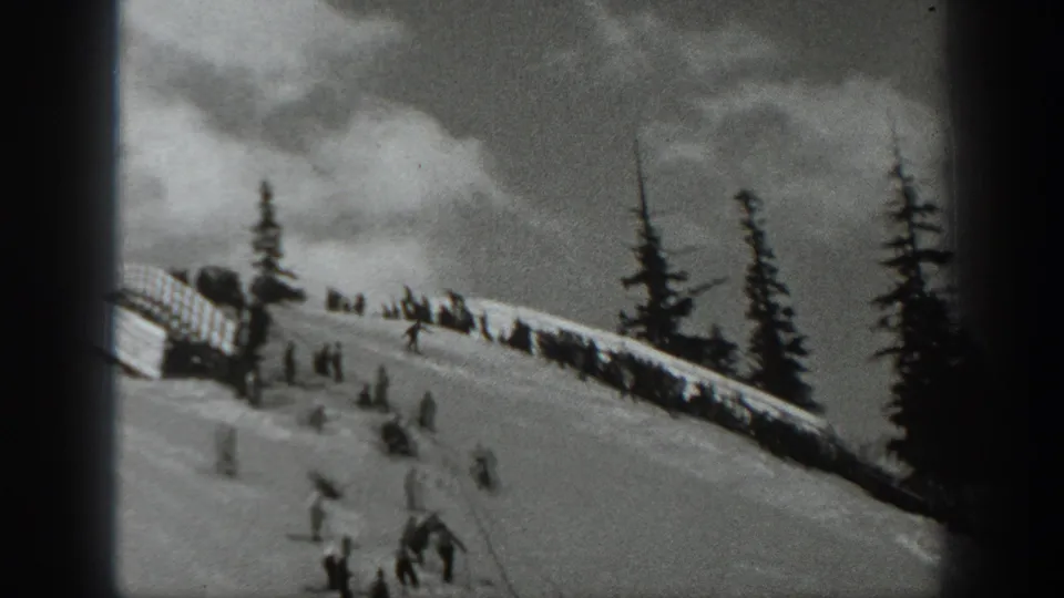 1940 Washington 8mm frame showing several distant skiers on a snowy slope beneath evergreens and a dark ridge or fence line.